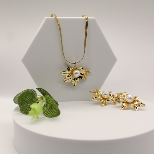 Gold-tone floral pendant necklace and matching flower-shaped earrings with white pearl centers, displayed on a white geometric stand with a small green plant prop against a light gray studio background.
