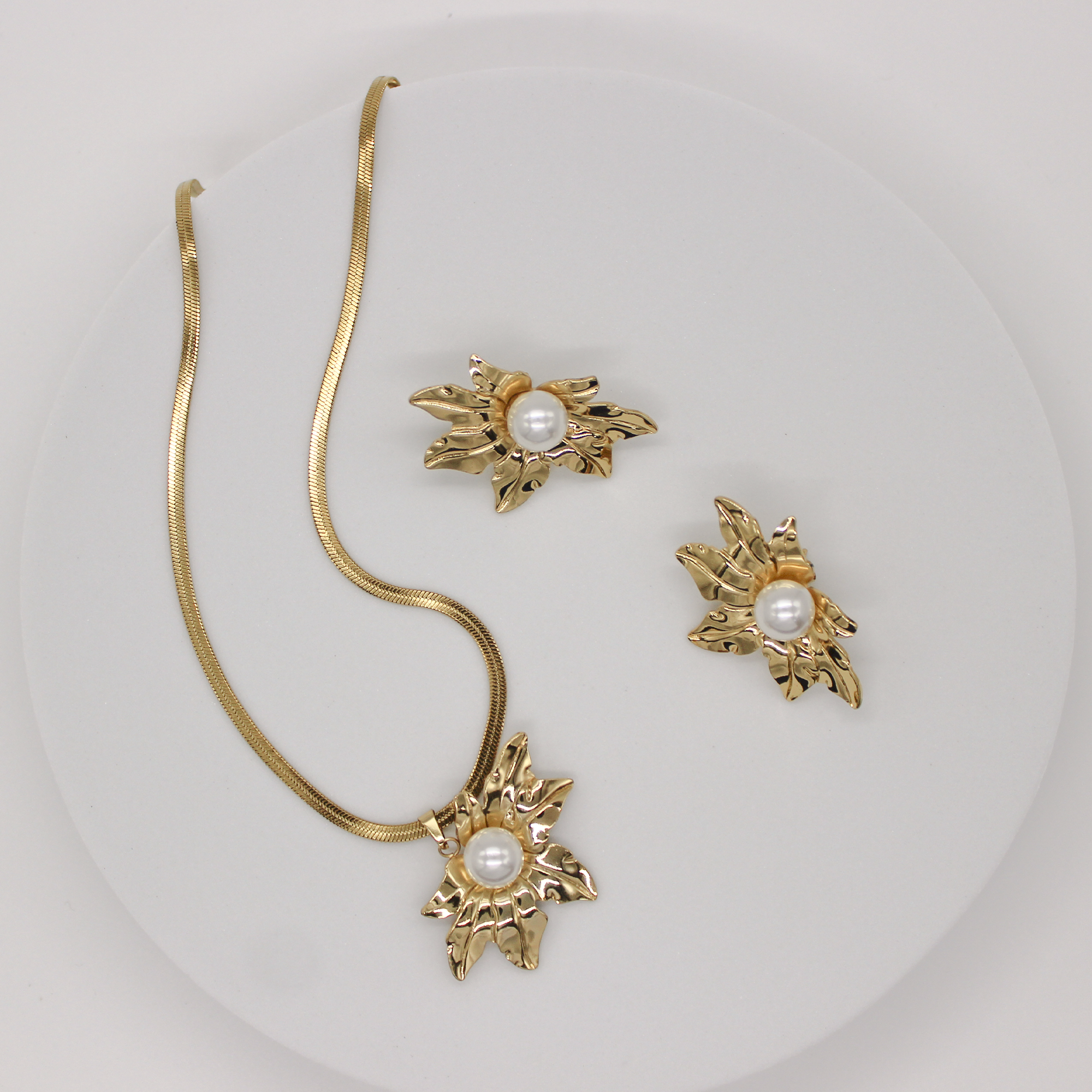 Gold jewelry set featuring a snake chain necklace and matching stud earrings, each decorated with a textured leaf-like gold motif surrounding a round white pearl, displayed on a white circular stand.