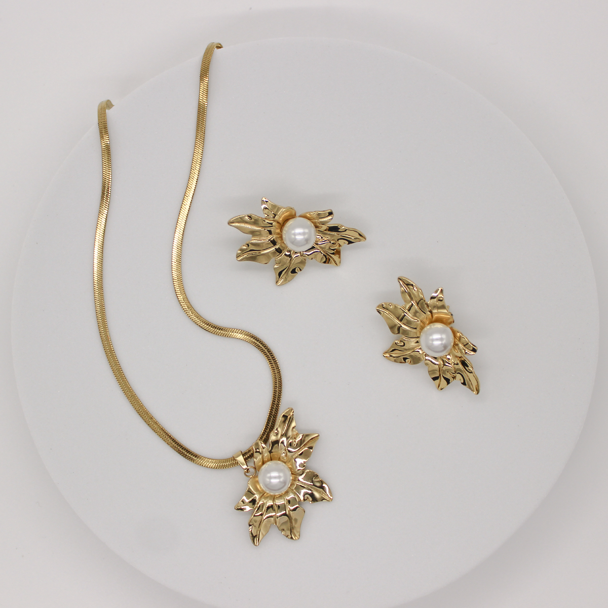 Gold jewelry set featuring a snake chain necklace and matching stud earrings, each decorated with a textured leaf-like gold motif surrounding a round white pearl, displayed on a white circular stand.