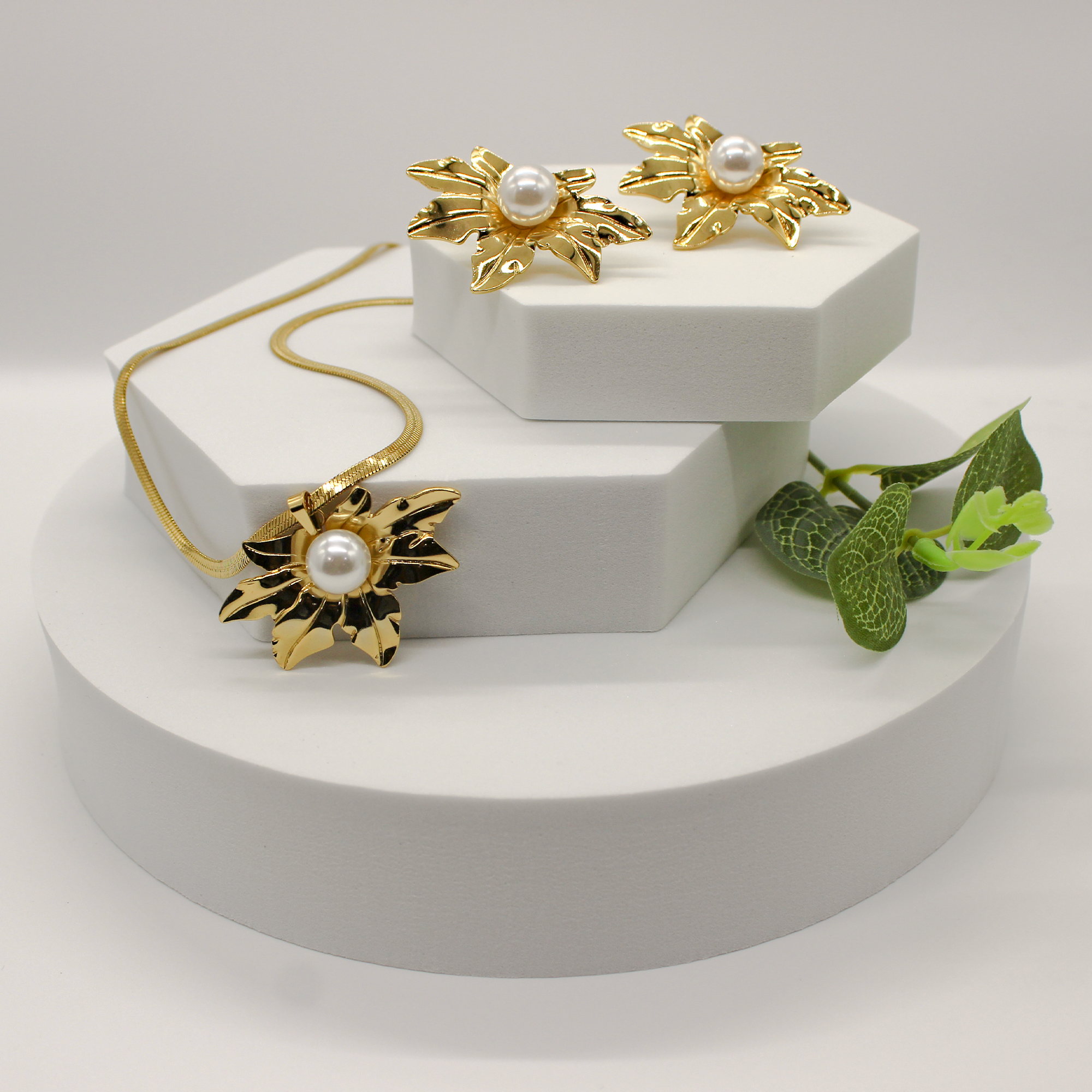 A gold jewelry set displayed on white geometric pedestals, featuring a necklace, a pair of earrings, and a decorative green leaf prop against a soft light gray background.