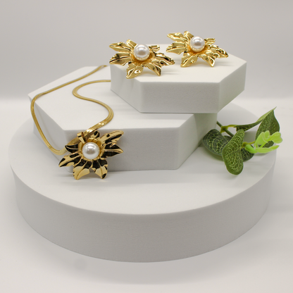 A gold jewelry set displayed on white geometric pedestals, featuring a necklace, a pair of earrings, and a decorative green leaf prop against a soft light gray background.