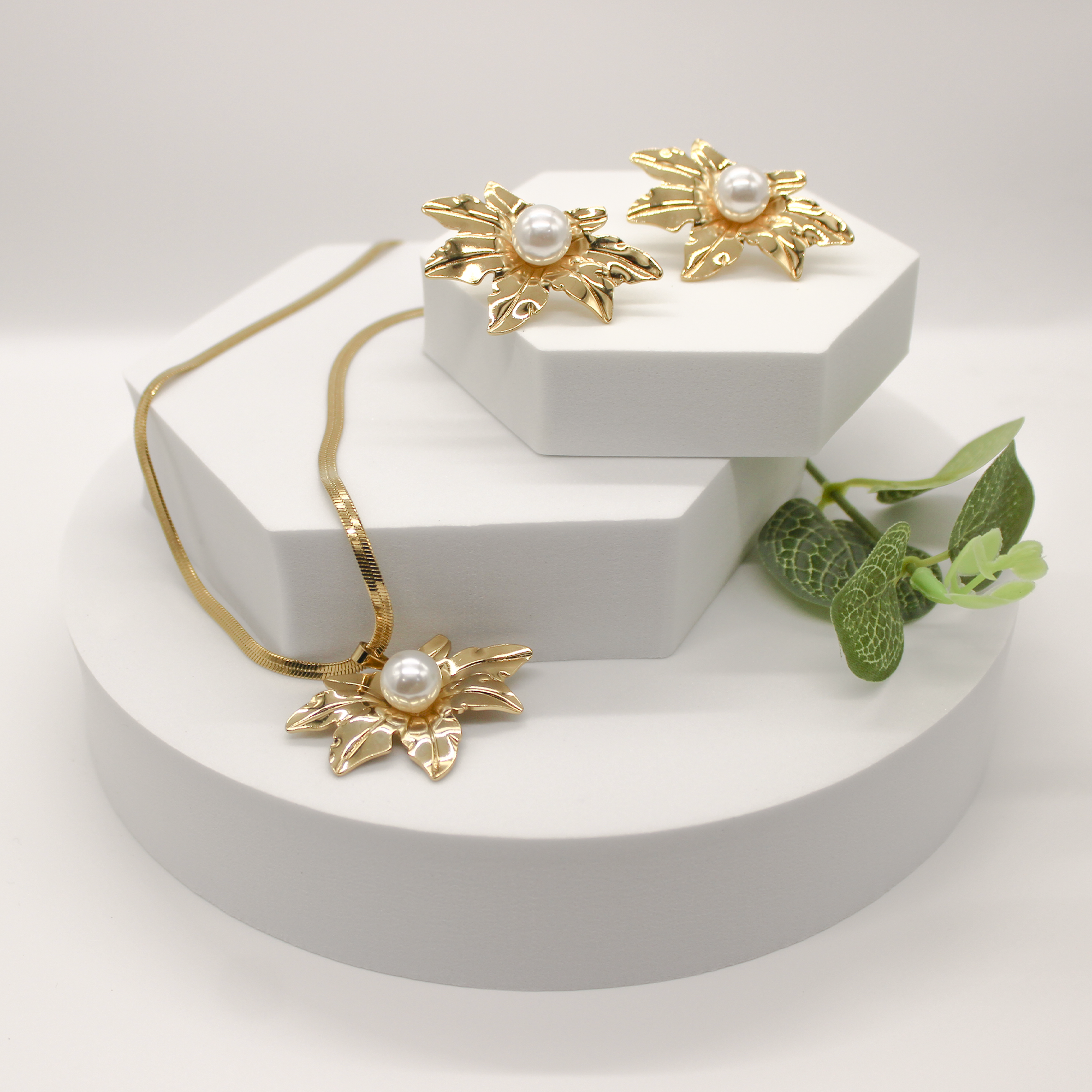 Gold floral jewelry set with a pendant necklace, matching earrings, and a green leafy sprig, displayed on white geometric blocks against a light gray background.